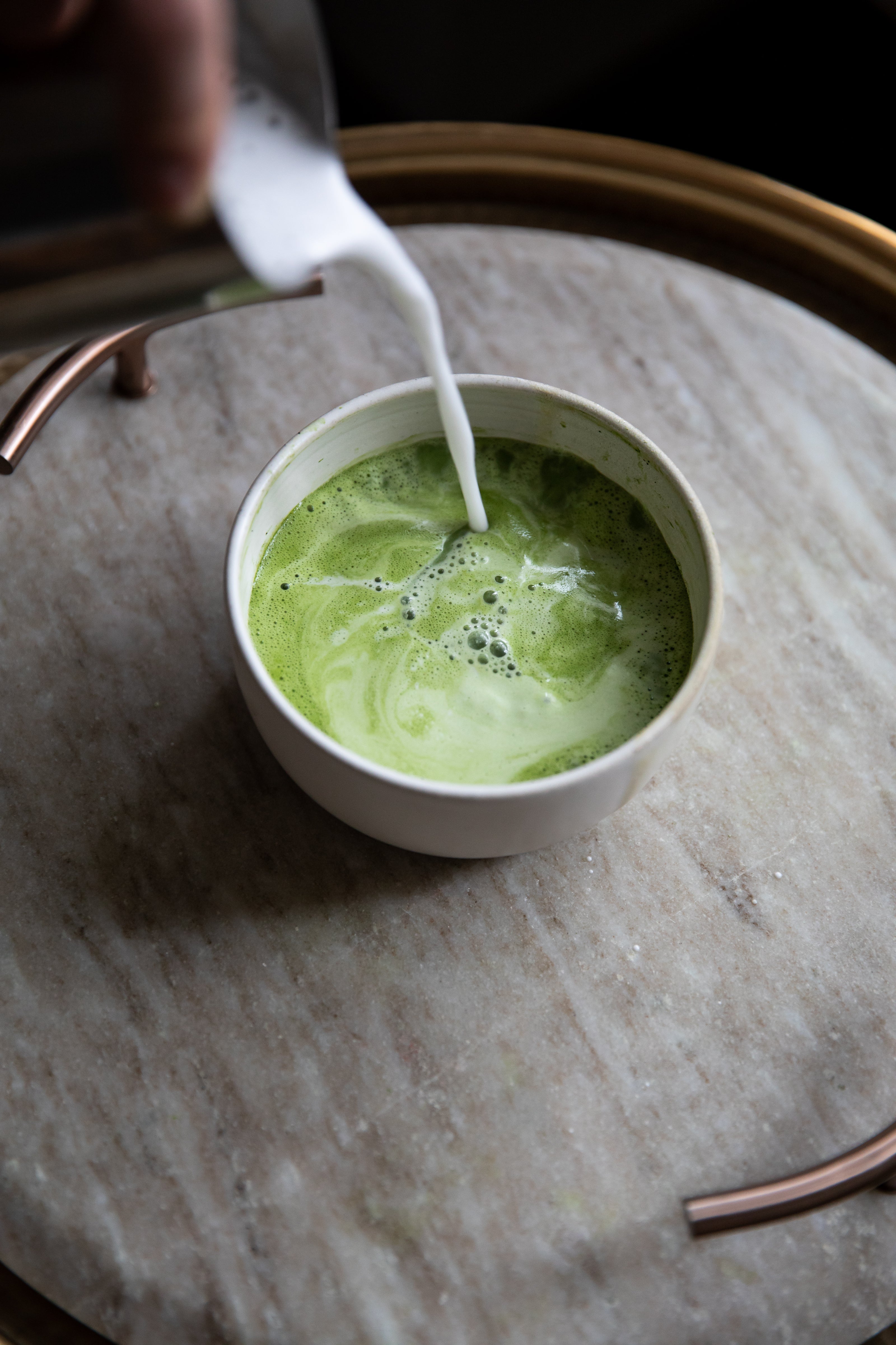 Green matcha latte being poured into a white cup on a marble surface.