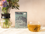 Tea box labeled 'Path of Clarity' with a glass of tea and flowers on a table.