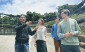 Four people standing outdoors on a paved path in front of a large agricultural structure with greenery and sky behind them. One man in a black jacket is gesturing while the others listen, and one person holds a small green leaf.