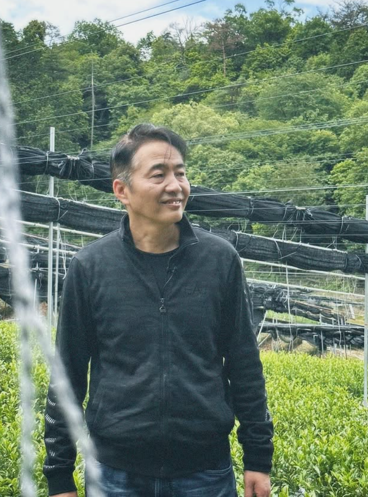 A smiling man wearing a black zip-up fleece jacket and blue jeans standing outdoors in a green agricultural field with rows of black tubers or plants behind him.
